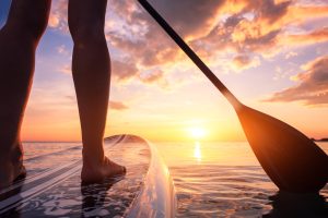 Stand up paddle boarding or standup paddleboarding on quiet sea at sunset with beautiful colors during warm summer beach vacation holiday, active woman, close-up of water surface, legs and board